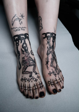 Rohes, handgezeichnetes Tattoo von Jette mit figürlichen Motiven, Strichzeichen und kreuzartigen Elementen über Füßen und Knöcheln.