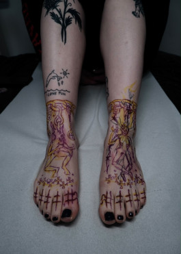 Expressive Tattoo-Arbeit von Jette mit roh gezeichneten Figuren und grafischen Zeichen in Schwarz sowie farbigen Akzenten auf beiden Füßen.